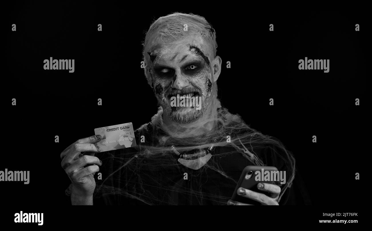 Sinister man with horrible scary Halloween zombie make-up using credit bank card and smartphone while transferring money, purchases online shopping. Dead guy with wounded bloody scars face, black room Stock Photo