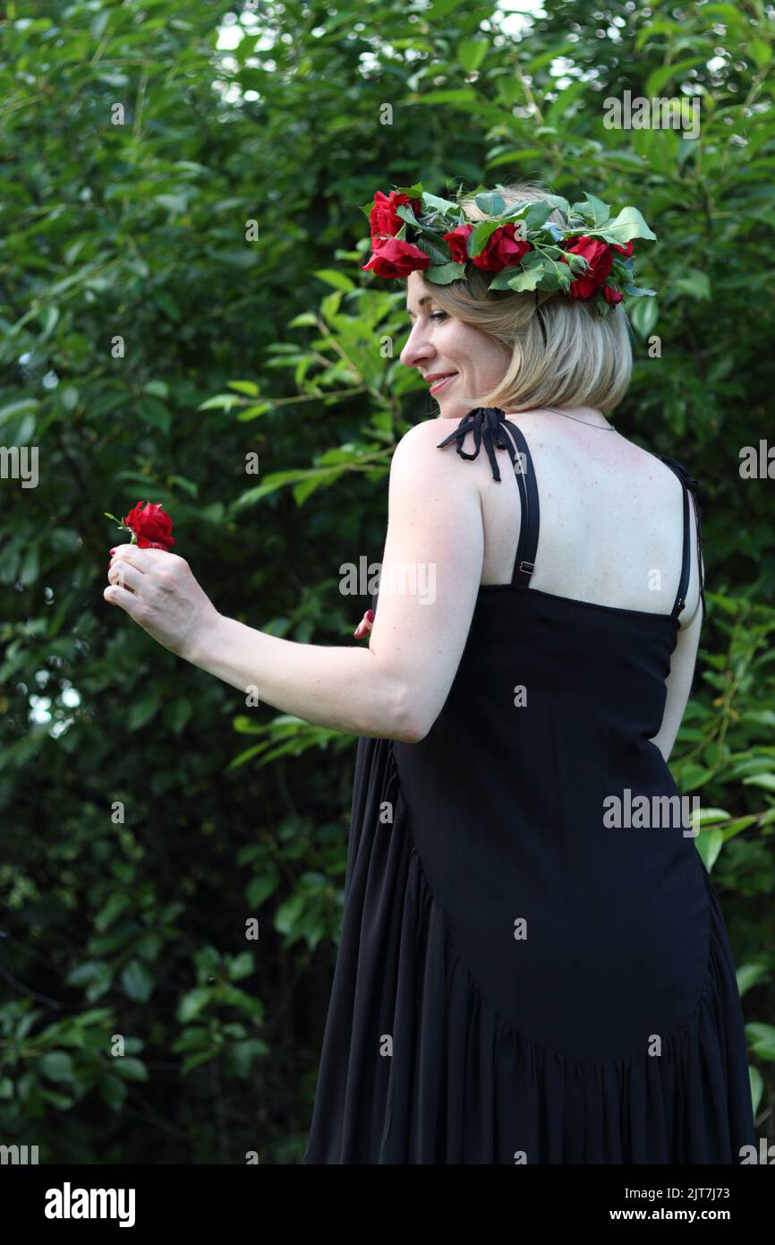 a standing young blonde woman with a short haircut in a black long dress and a red wreath with a red flower in front of green trees, side face Stock Photo