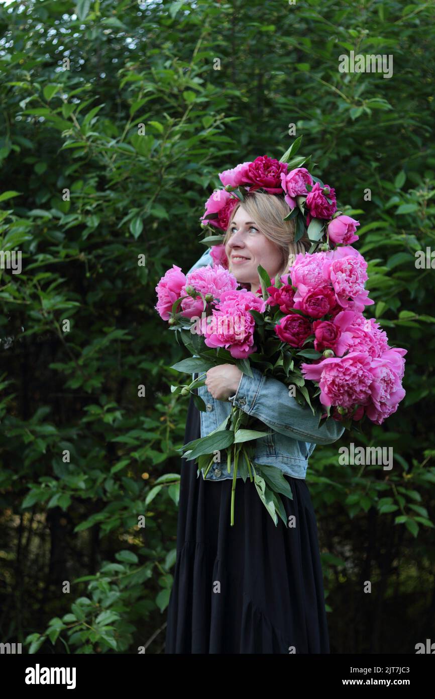 a standing young blonde woman in wreath holding a bouquet of pink flowers in front of green trees in the summer, looking up in the sky, side face Stock Photo