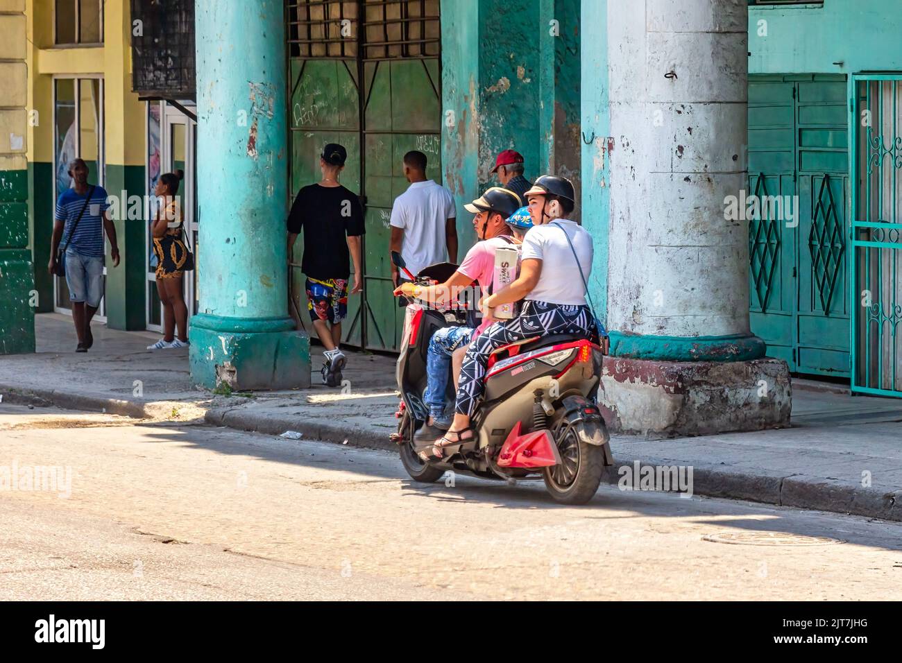 An electric bicycle carries a family of three people. Other people are walking in a porch where buildings are weathered and dirty Stock Photo