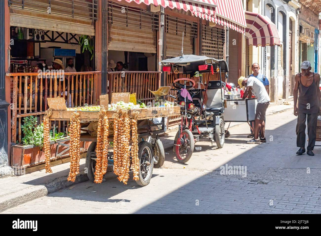 A food selling cart is parked in a narrow city street in Old Havana. Other small businesses are in the background. A slim man walks in the area. Stock Photo