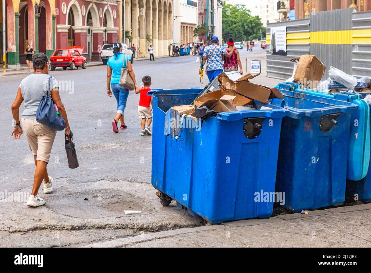 Cuban people walking by large garbage bins in a city street. The waste is uncovered and overflowing. Stock Photo