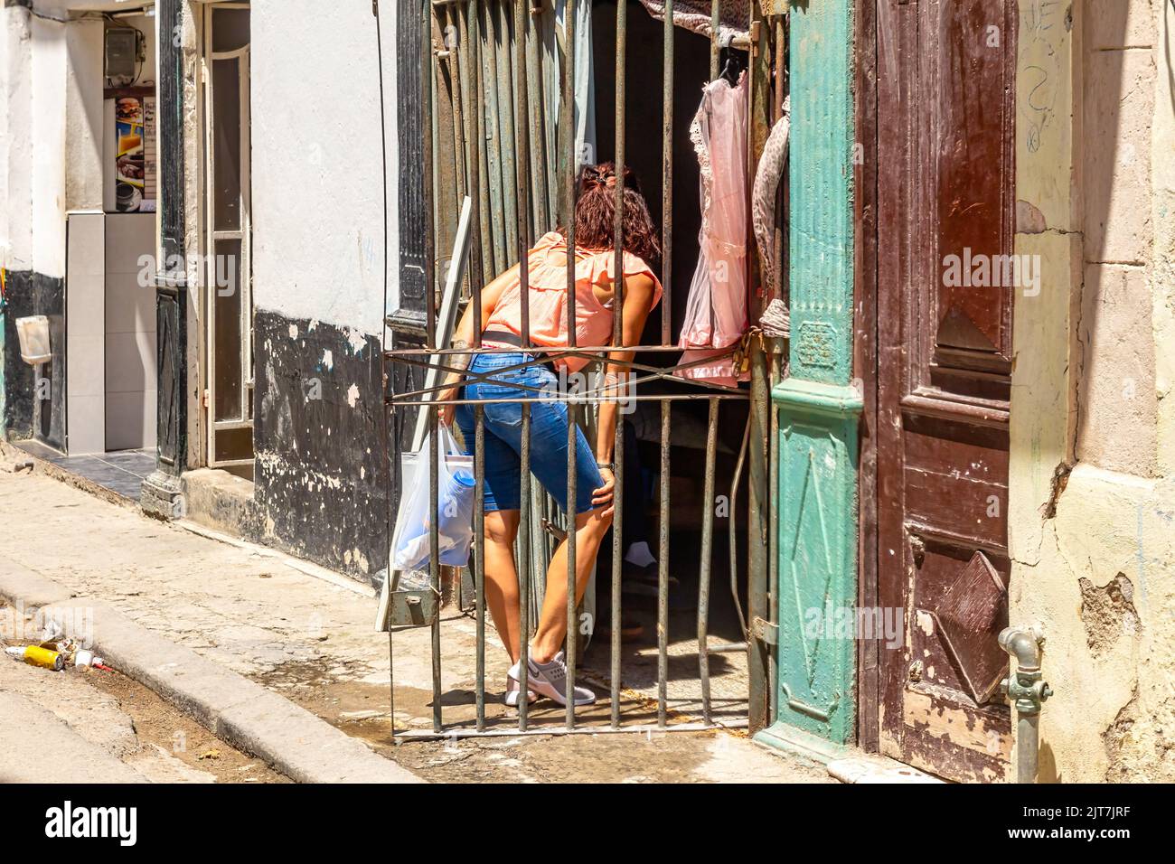 A Cuban young woman stands in front of a house door which is protected by a metalwork. The sidewalk dirty and broken Stock Photo
