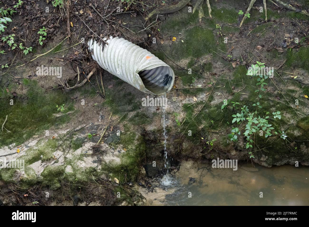 A corrugated metal drain with a light trickle of water draining into a small creek Stock Photo
