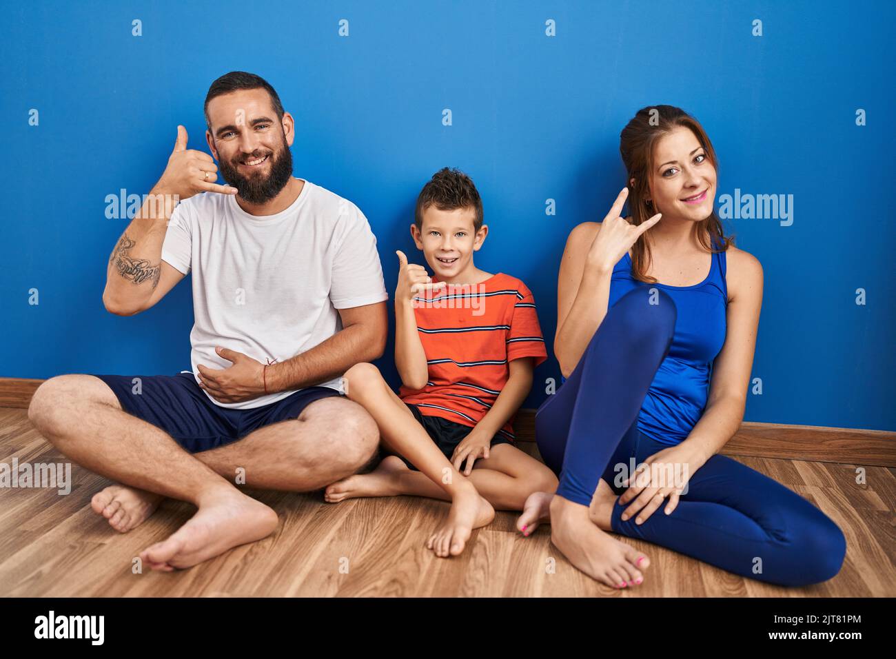 Family of three sitting on the floor at home smiling doing phone gesture with hand and fingers like talking on the telephone. communicating concepts. Stock Photo