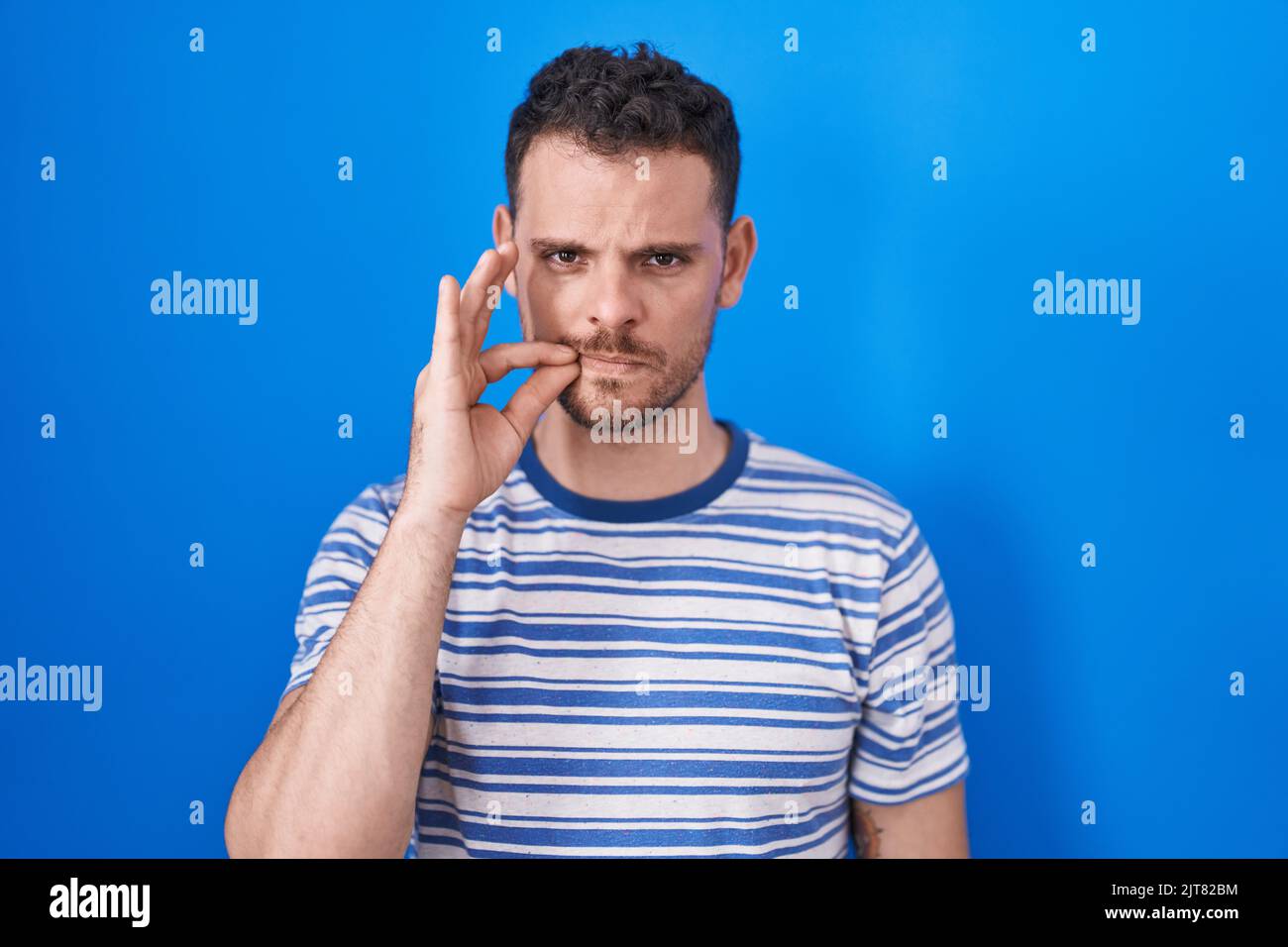 Young hispanic man standing over blue background mouth and lips shut as ...