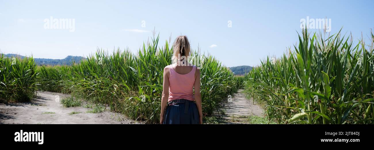 Wide view image of a young woman standinh in the middle of green corn fields on a crossroads deciding which way to go. Stock Photo