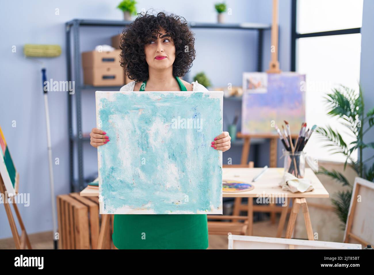 Young brunette woman with curly hair holding painter canvas smiling looking to the side and staring away thinking. Stock Photo