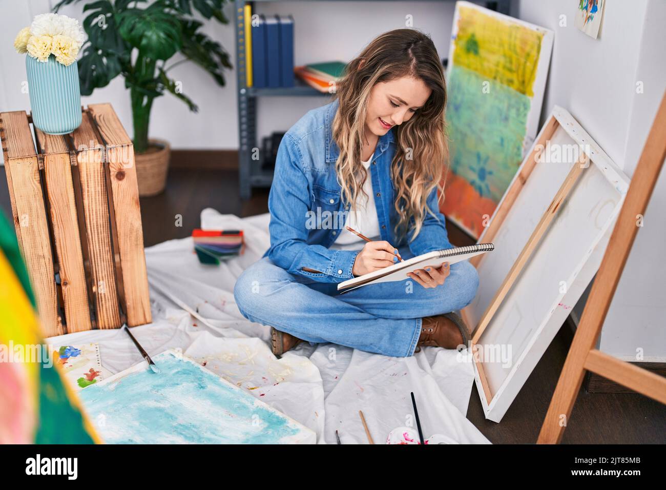 Young woman artist drawing on notebook sitting on floor at art studio ...