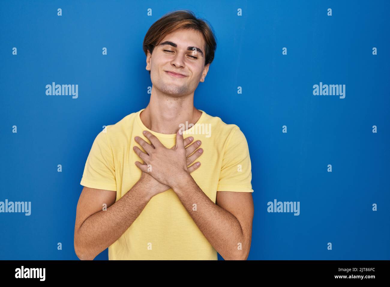 Young man standing over blue background smiling with hands on chest ...