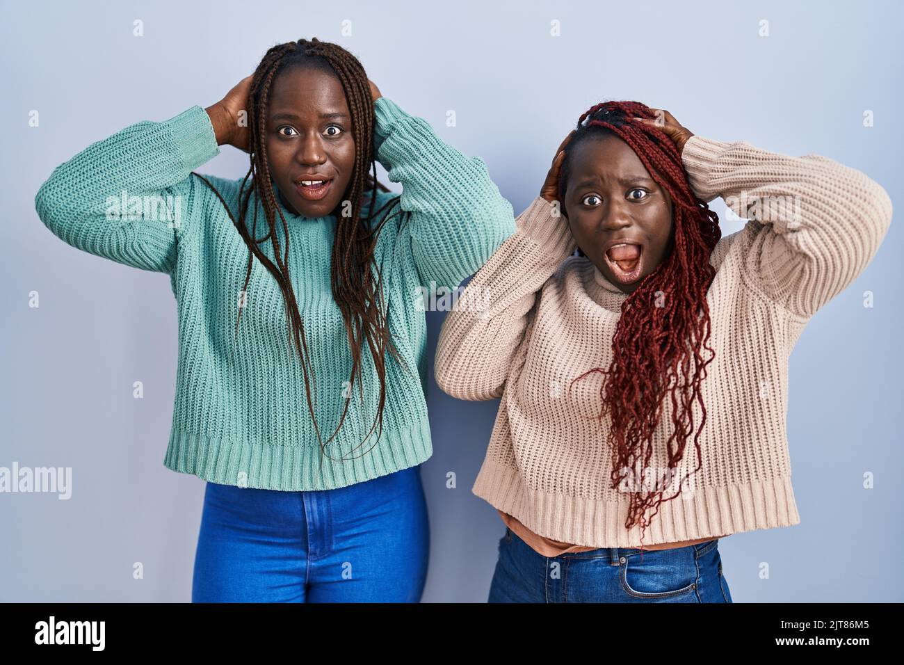 Two african woman standing over blue background crazy and scared with hands on head, afraid and surprised of shock with open mouth Stock Photo