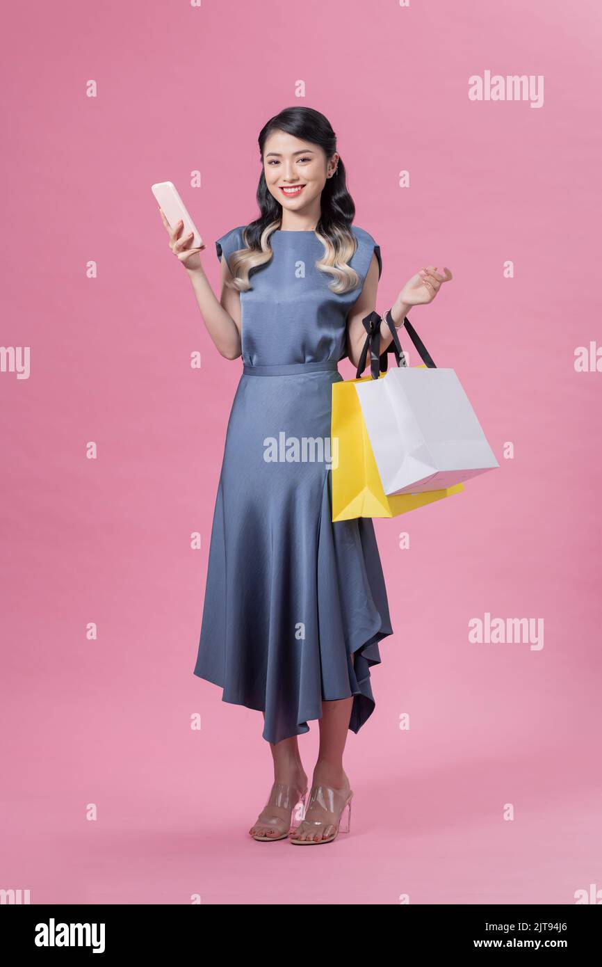 Cheerful excited asian woman using mobile phone carrying shopping bags full body standing on pink background. Stock Photo