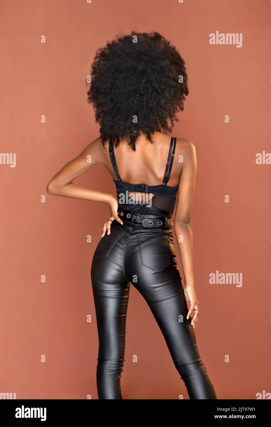 Back view of anonymous African American female model with Afro hairstyle in leather pants and top standing with hand on waist against brown background Stock Photo