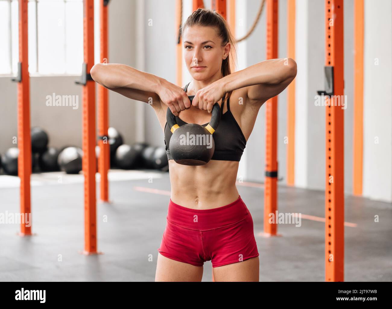 Strong female athlete in bra and shorts doing kettlebell high pull exercise and looking away during functional workout on blurred background of light Stock Photo