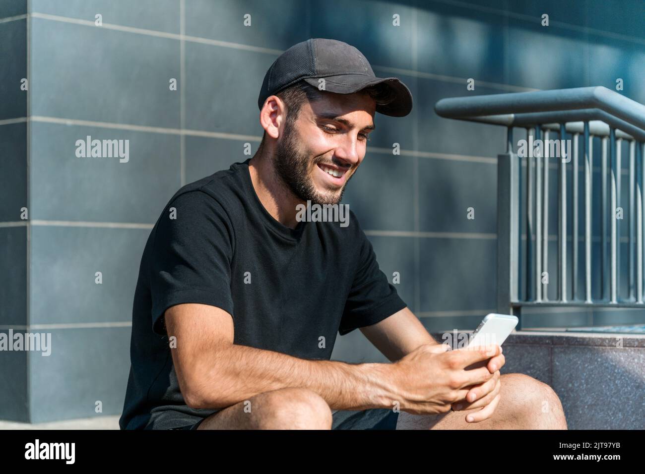 Cheerful male in casual clothes and cap smiling and sending text message in social media on cellphone while sitting on steps outside modern building o Stock Photo