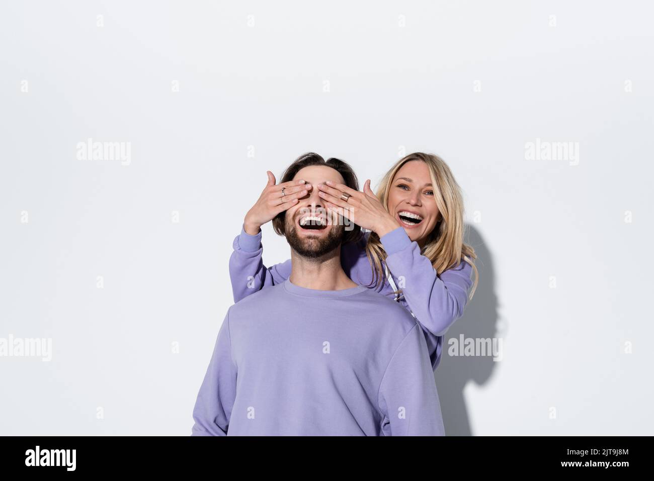 happy blonde woman covering eyes of bearded man in purple sweatshirt on grey,stock image Stock Photo
