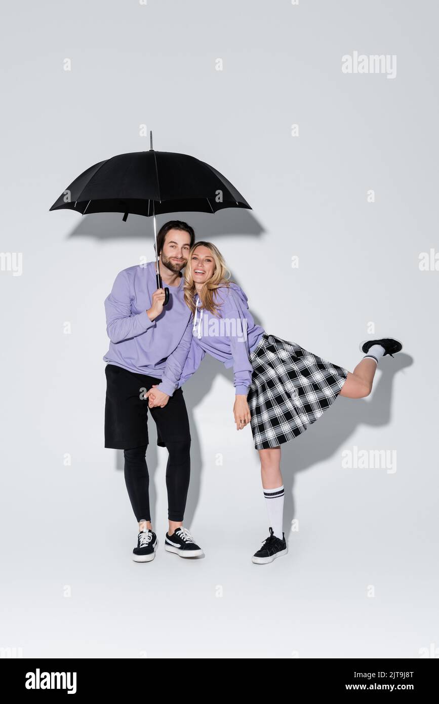 full length of happy couple in purple sweatshirts standing under umbrella on grey,stock image Stock Photo