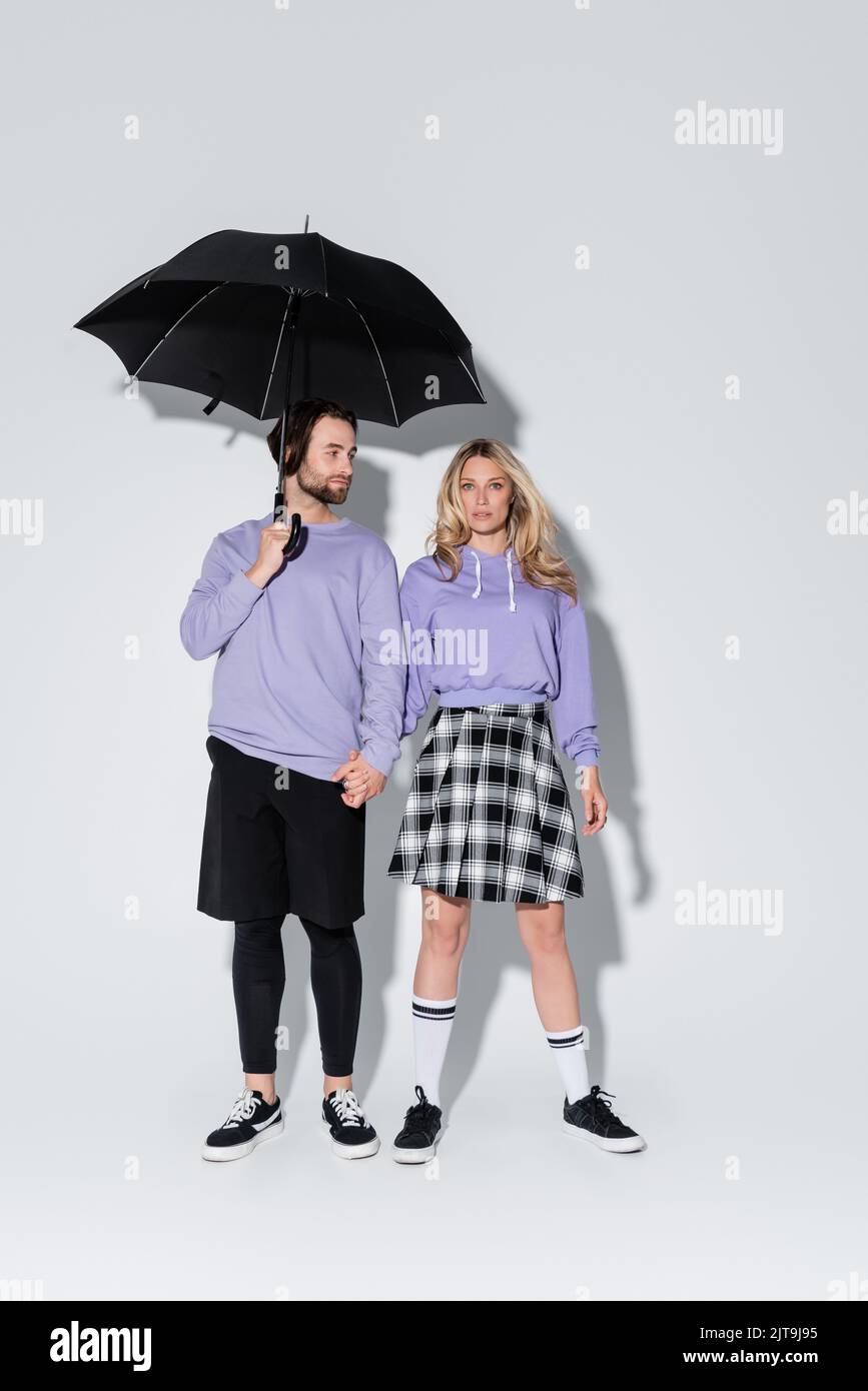 full length of stylish couple in purple sweatshirts standing under black umbrella on grey,stock image Stock Photo