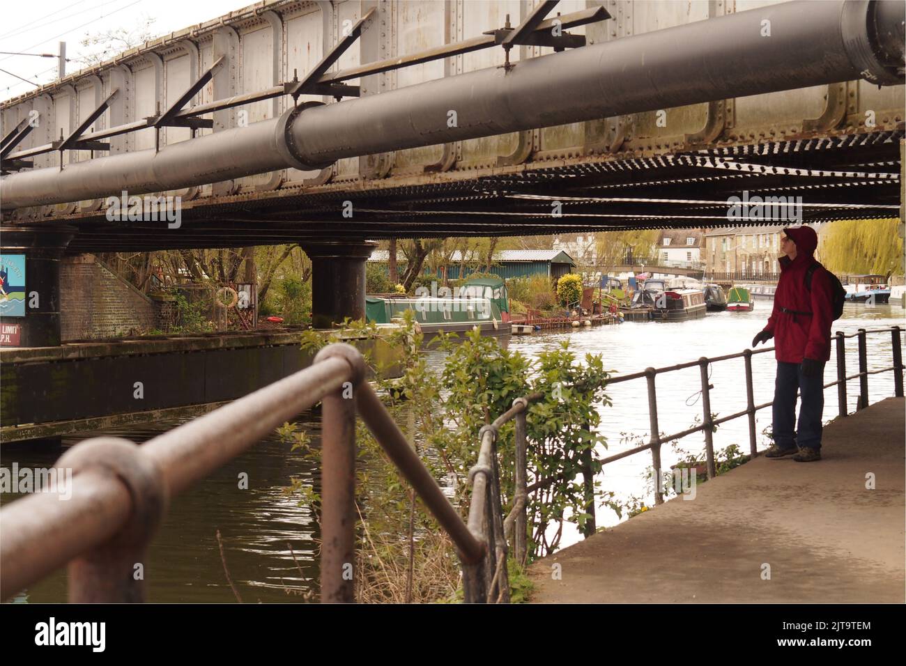 A man 60+ standing under a railway bridge on a canal tow path at Ely, Cambridgeshire, studying the underside of the bridge wearing a waterproof coat Stock Photo