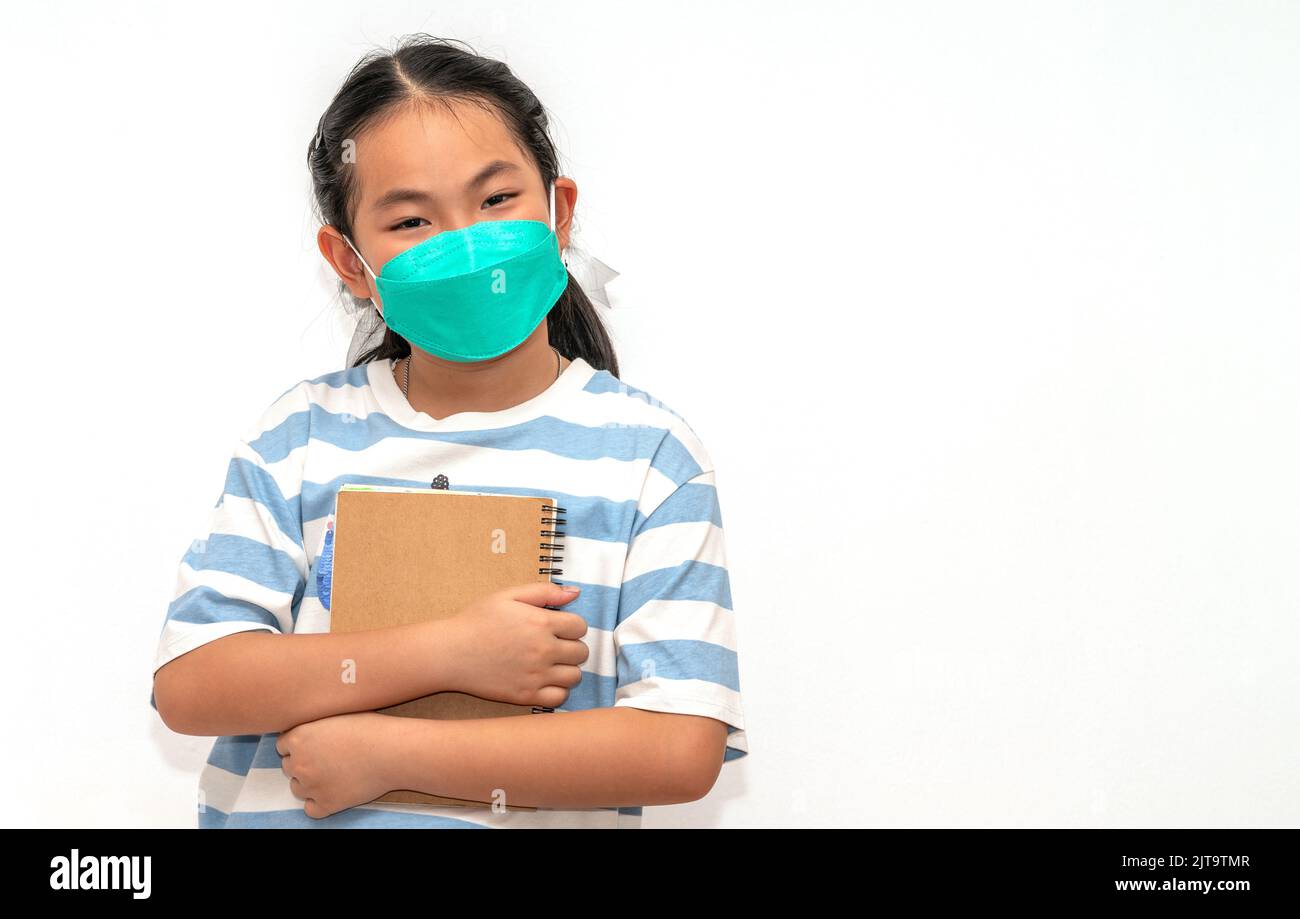 Cute student Asian child girl holding a book, wearing face mask for protection in era of covid-19, isolated student child girl on white background, co Stock Photo