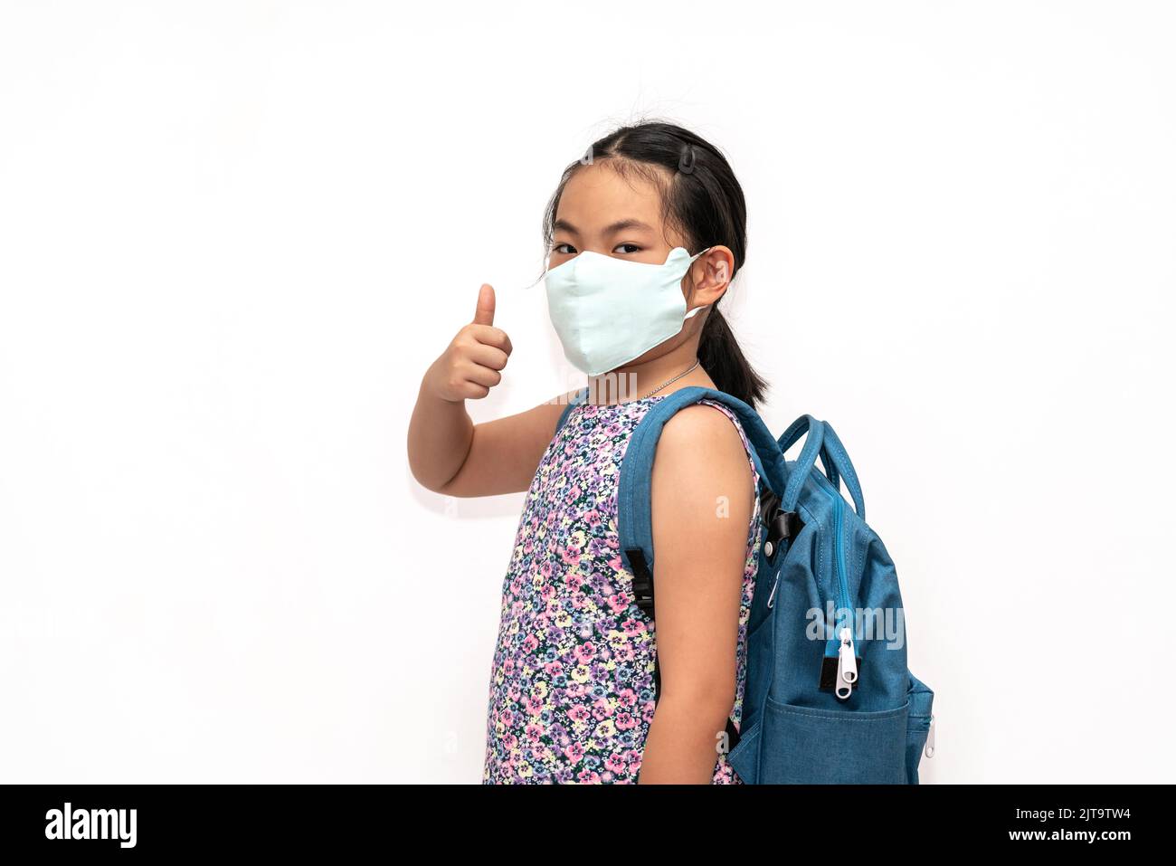 Portrait Asian child girl with facemask, carrying a blue backpack, show thumb up with confident, isolated portrait image on white background. The conc Stock Photo