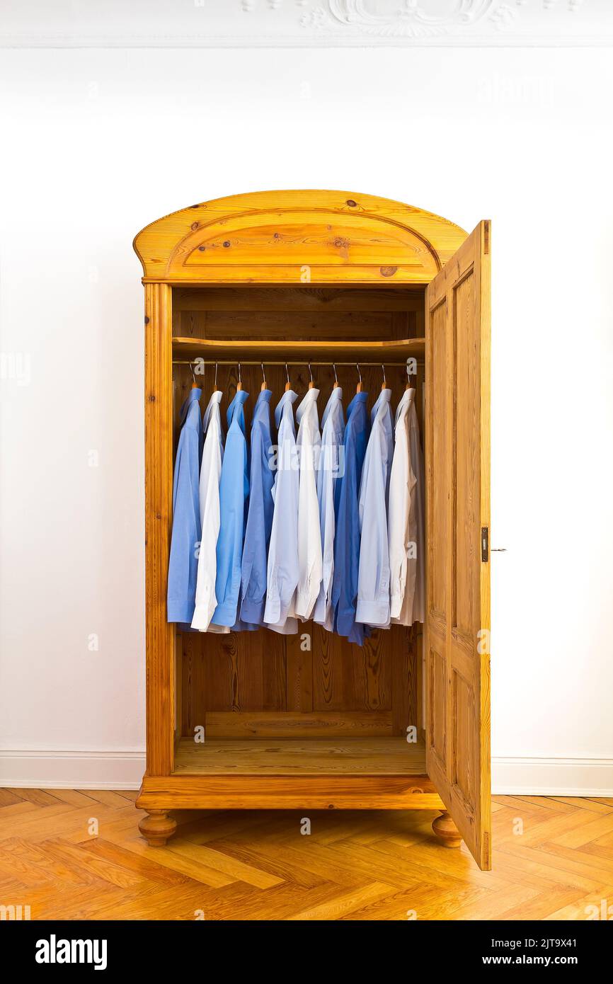 Open antique wardrobe with blue dress shirts, against the white wall of an old building with parquet flooring and stucco plastering, copy space. Stock Photo