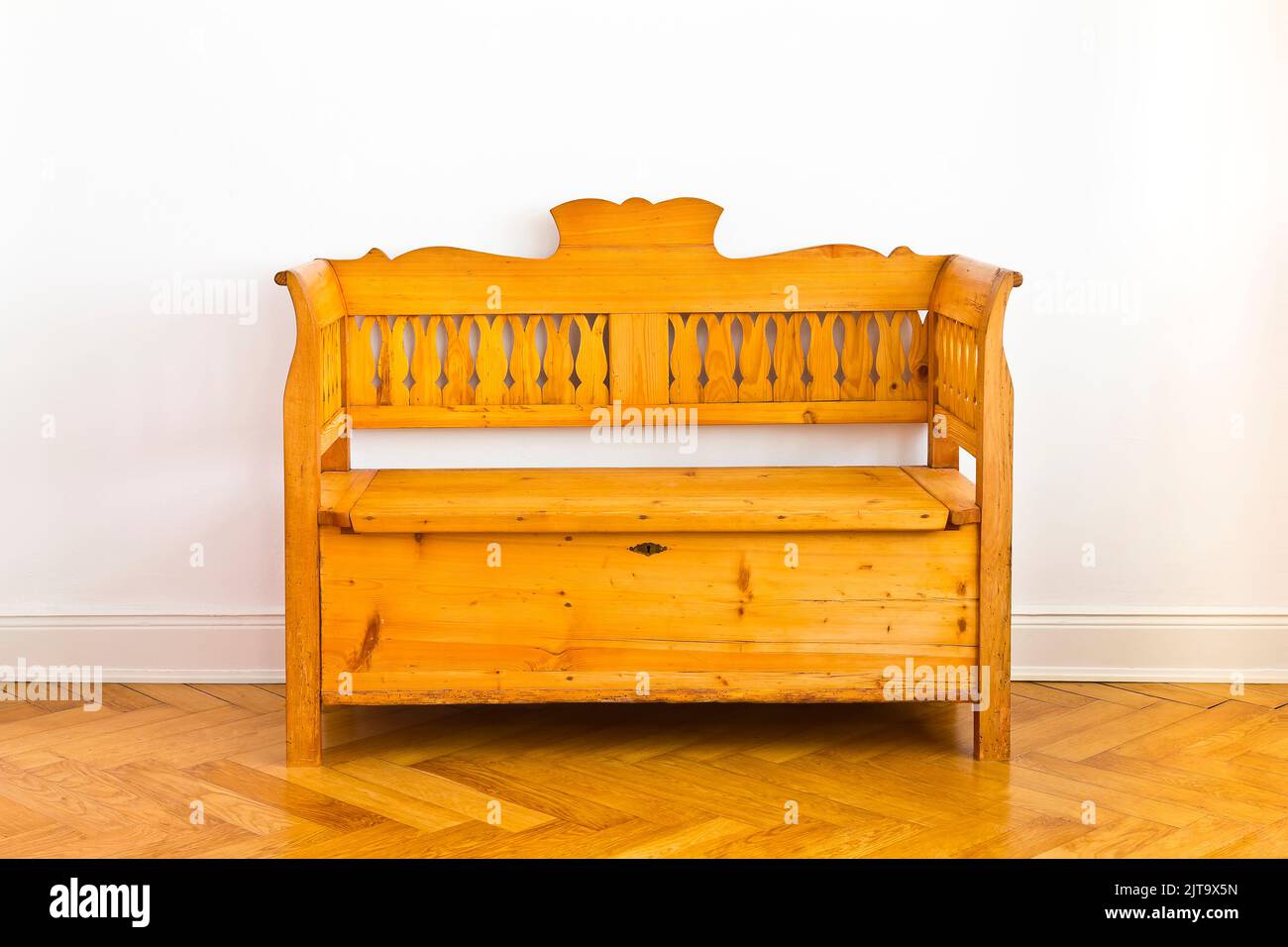 Restored antique wooden box bench with storage space unter the seat, circa 1880, against the white wall of an old building with parquet flooring. Stock Photo