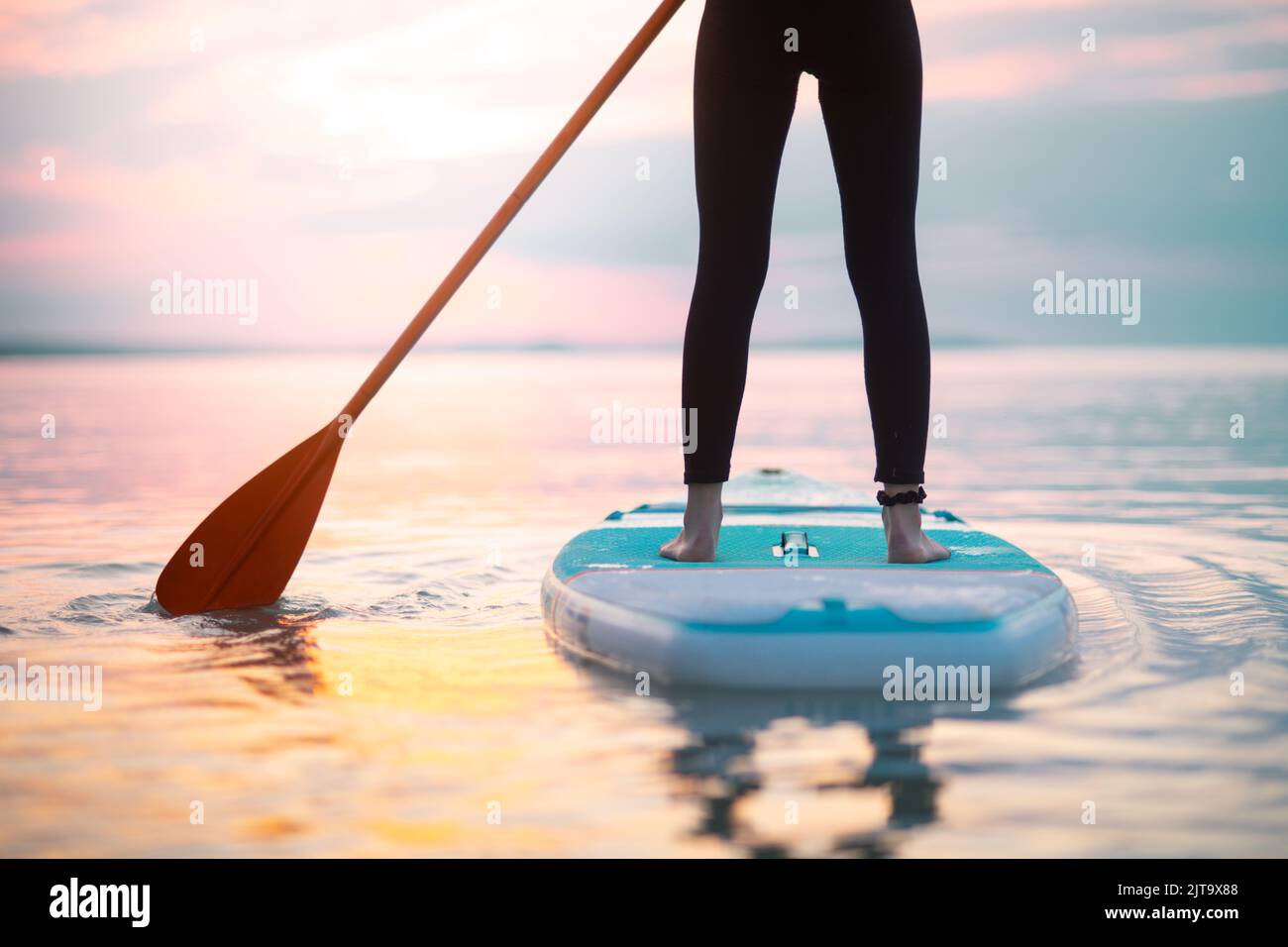 Rear view of girl surfer paddling on surfboard on the lake at sunrise, lowsection. Stock Photo