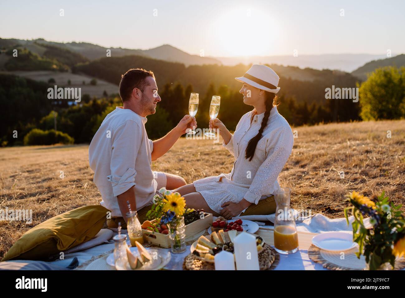 Attractive couple in love enjoying picnic and drinking wine on the hill at sunset. Stock Photo