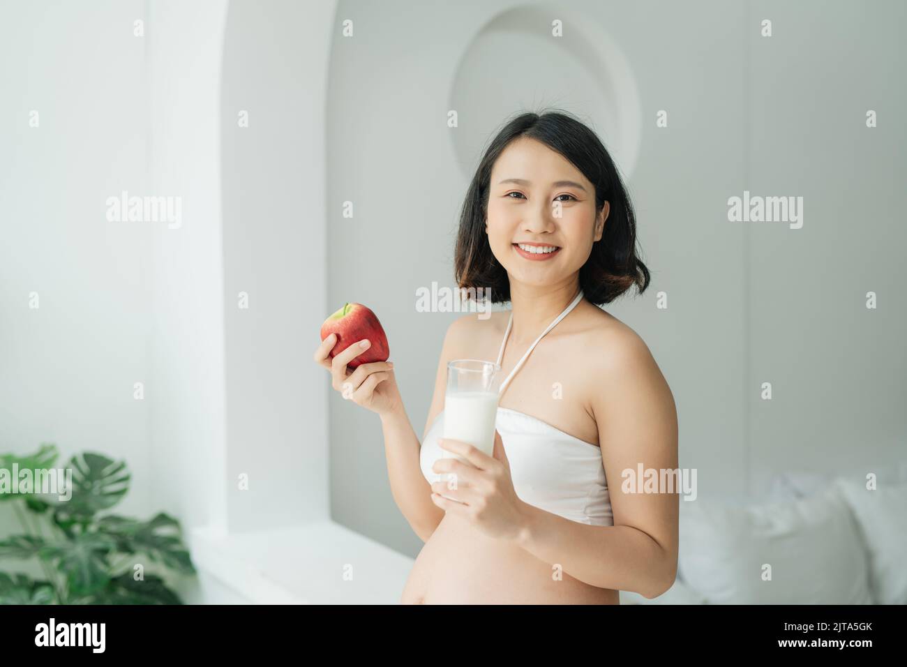Pregnant Asia woman holding a glass of milk and red apple at home Stock Photo