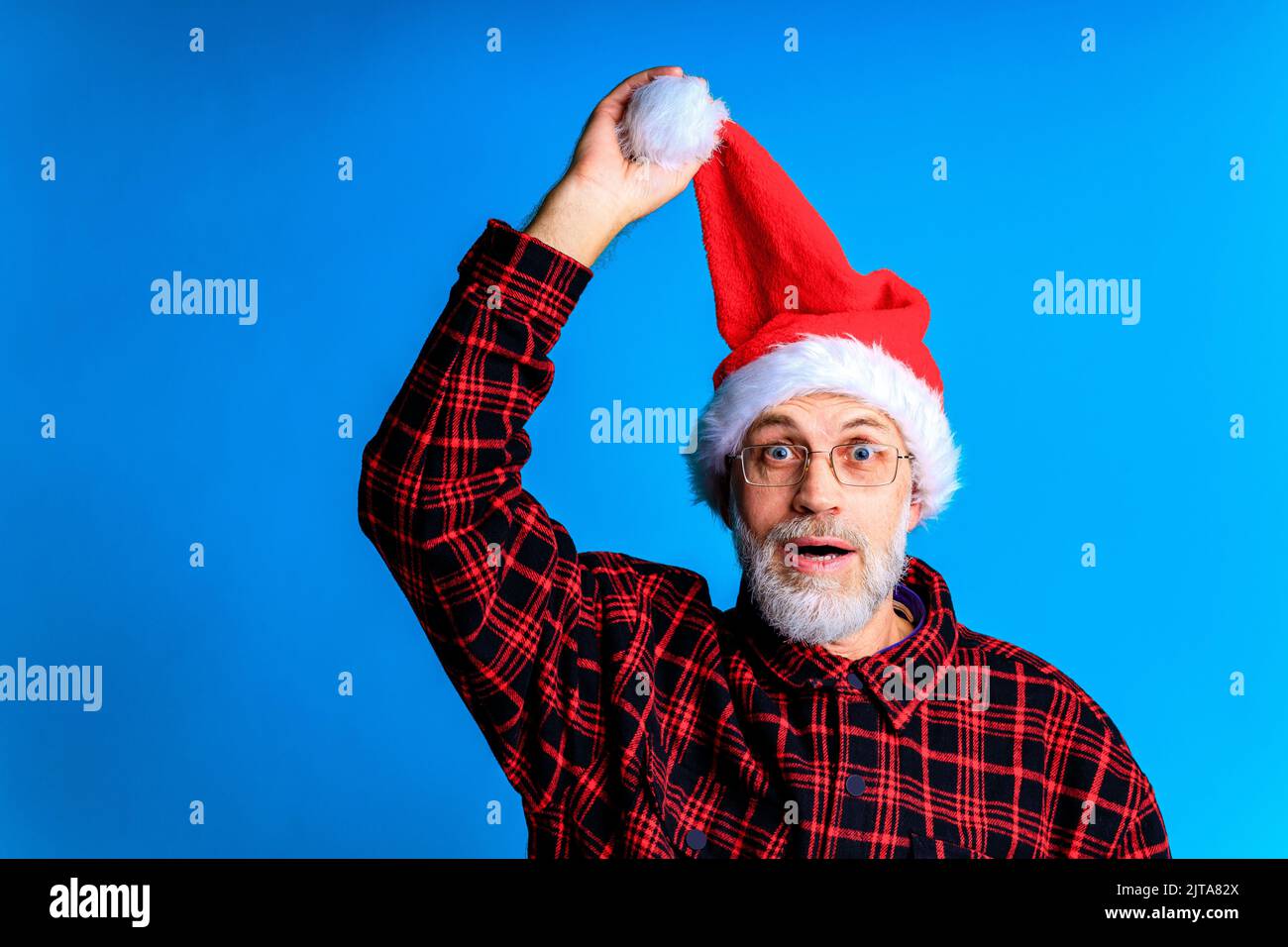 an elderly and well-groomed man in a plaid shirt and santa hat in blue studio background Stock Photo