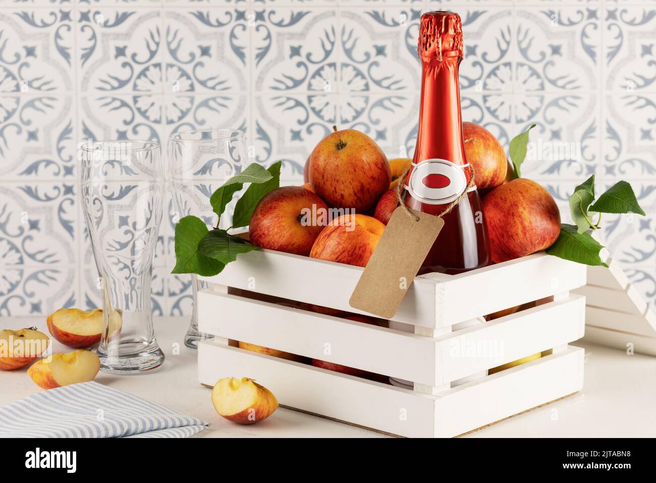 Apple cider drink or fermented fruit drink. Bottle with cider, glasses and red organic apples in a white wooden box on a kitchen table. Healthy eating Stock Photo