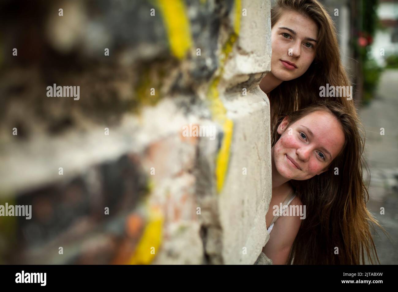 Two pretty girls look out from around the corner of a brick house. Stock Photo
