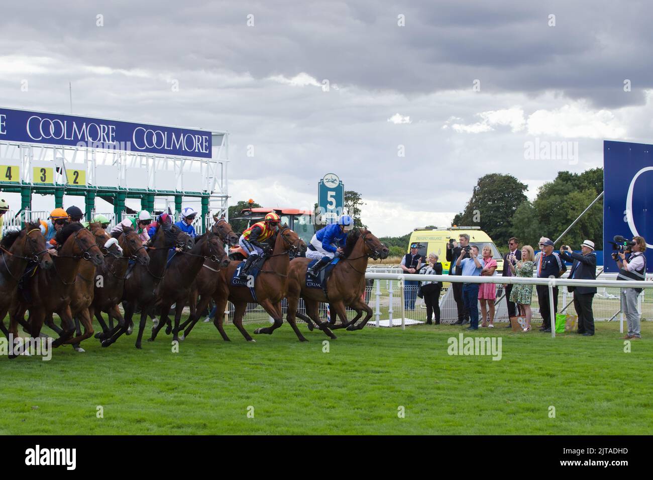 Jockeys and their horses leave the starting gates during the Coolmore Wootton Bassett Nunthorpe Stakes as spectators and a camera crew look on. Stock Photo