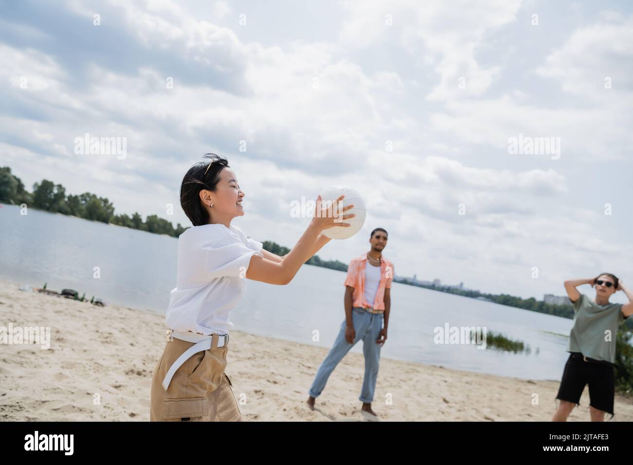 cheerful asian woman holding ball while playing beach volleyball with multiethnic friends Stock Photo