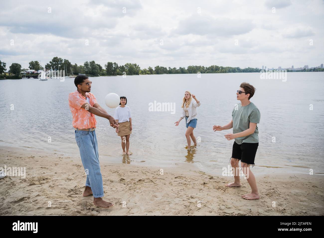 interracial men playing beach volleyball near women standing in water Stock Photo