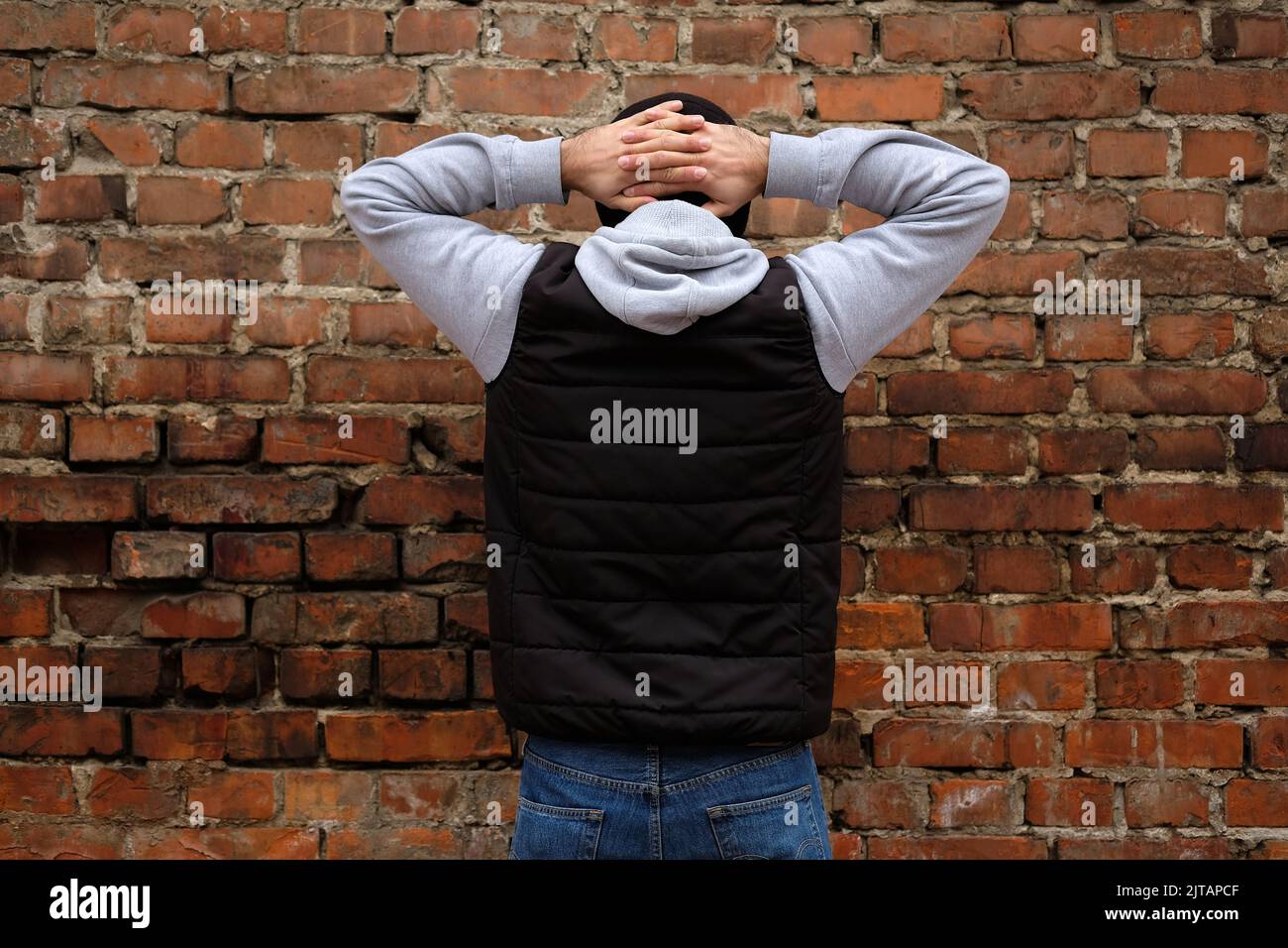 The guy stands facing a brick wall with his hands clasped behind his head. A guy in jeans and a black puffy tank top with his back to the camera. Arre Stock Photo