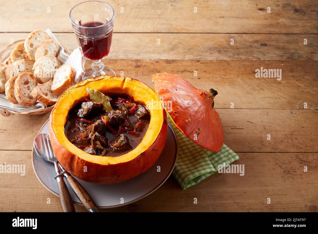 From above delicious meat goulash served in pumpkin near plate with bread pieces and glass of red wine on timber table during lunch Stock Photo