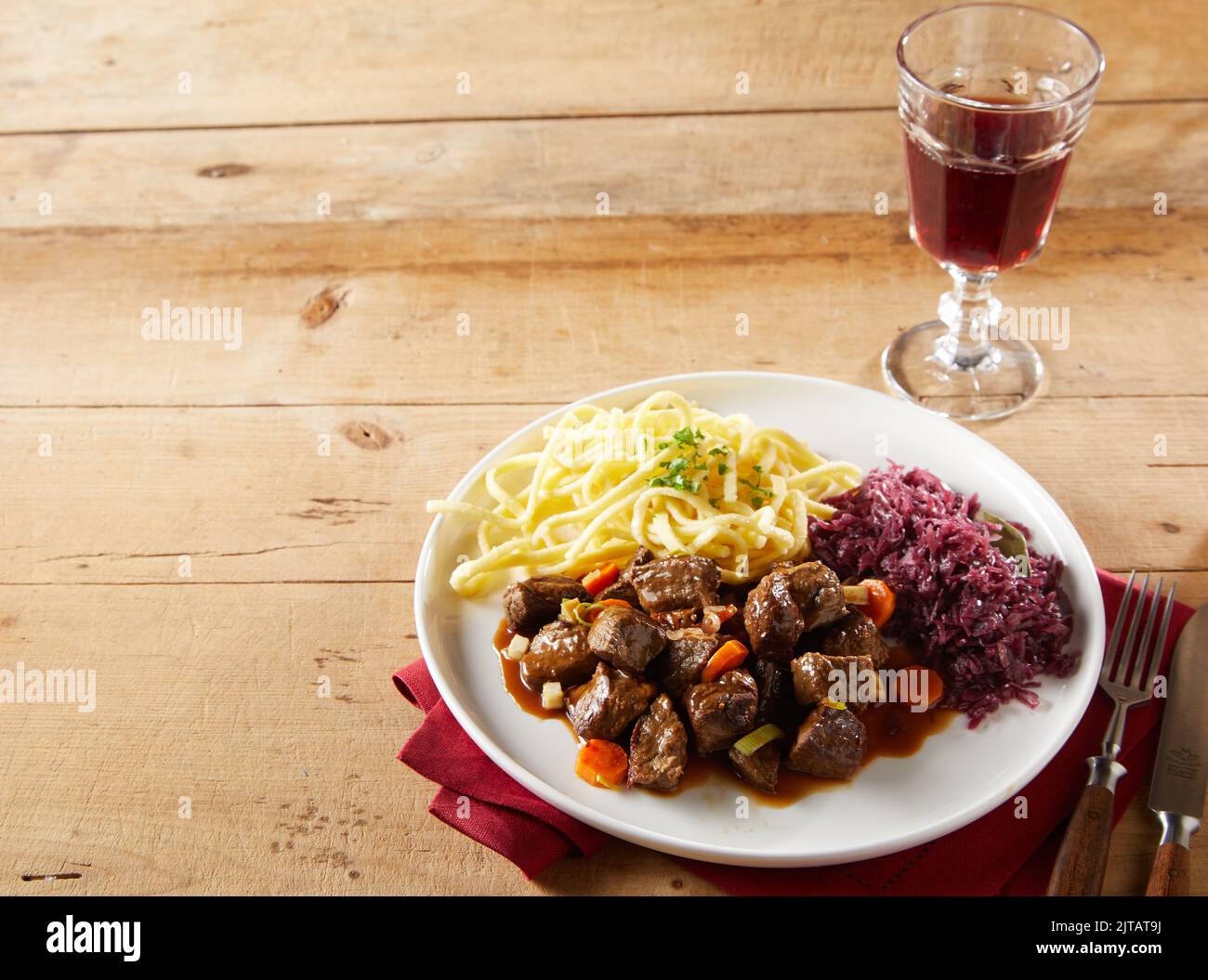 From above tasty beef goulash and spatzle pasta with sauerkraut served for lunch on plate near glass goblet of red wine on timber table in daytime Stock Photo