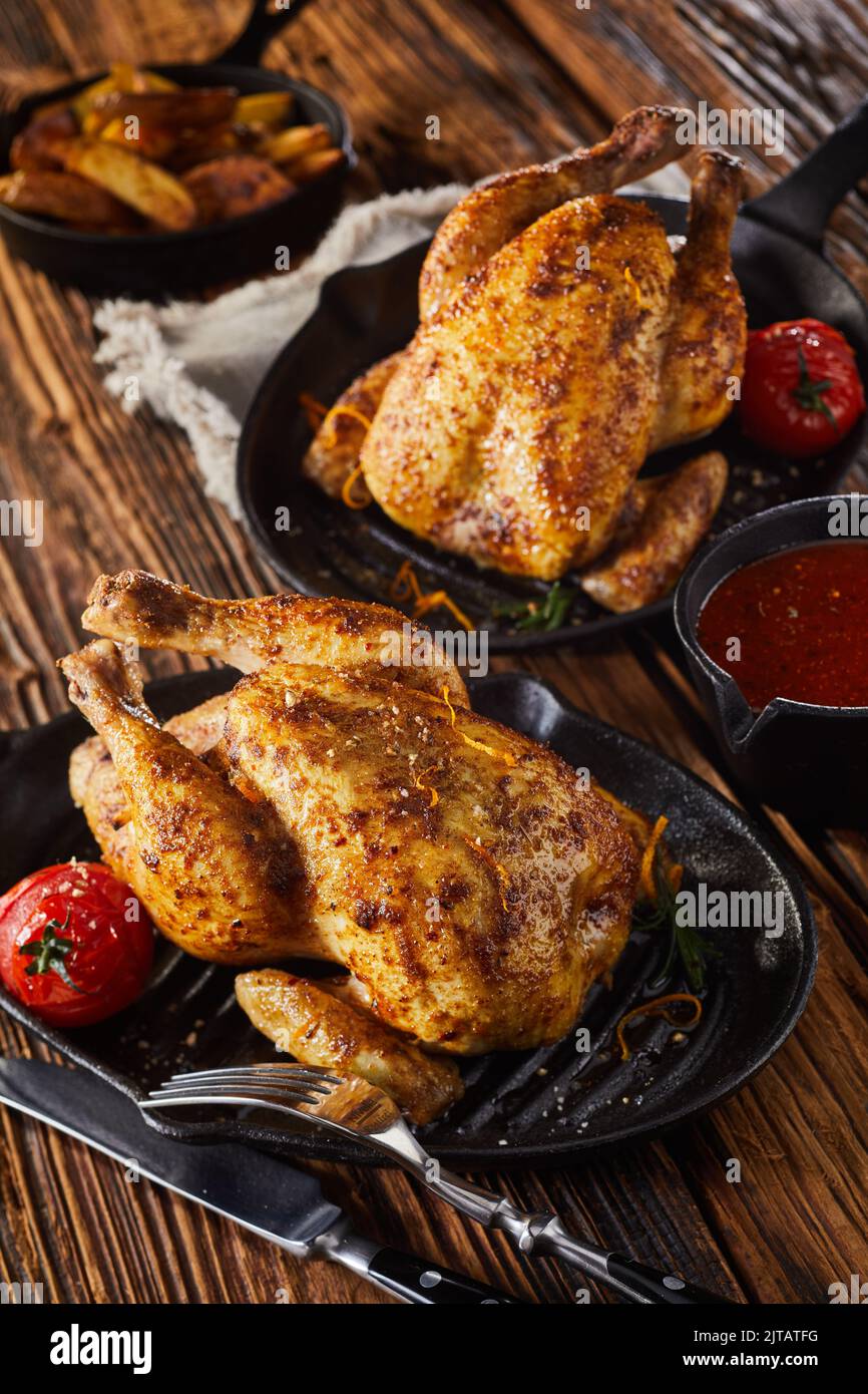 From above whole corn fed chickens roasted with tomatoes and served on frying pans near saucepan and cutlery on timber table Stock Photo