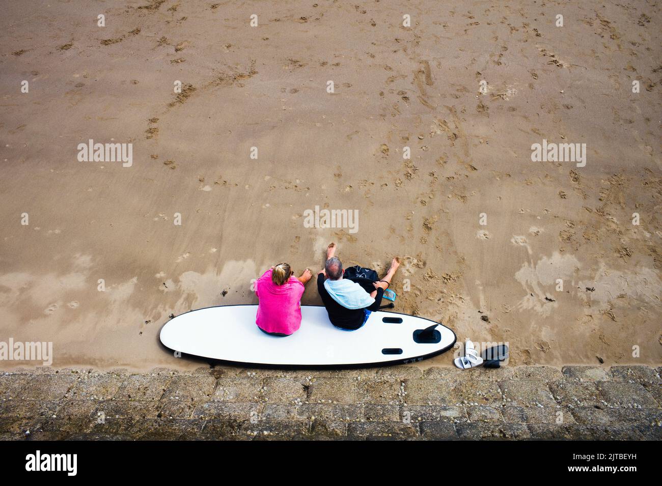 Looking down on an older couple sitting on a surfboard on a sandy beach Stock Photo