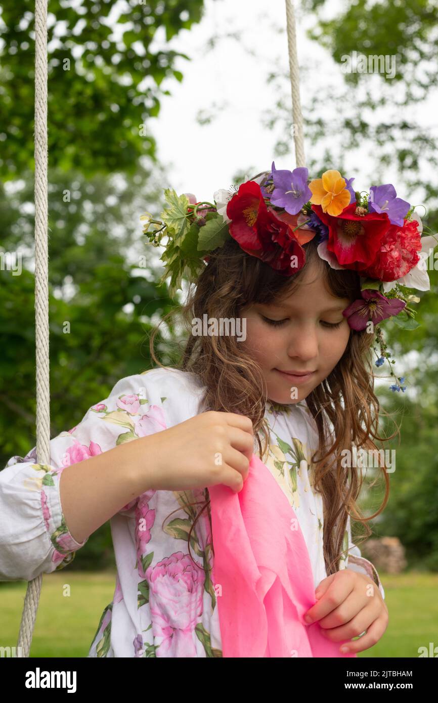 A contemplative girl with long hair wears a flower crown with red poppies, blue campanula, and leaves, while sitting on an outdoor swing. Stock Photo