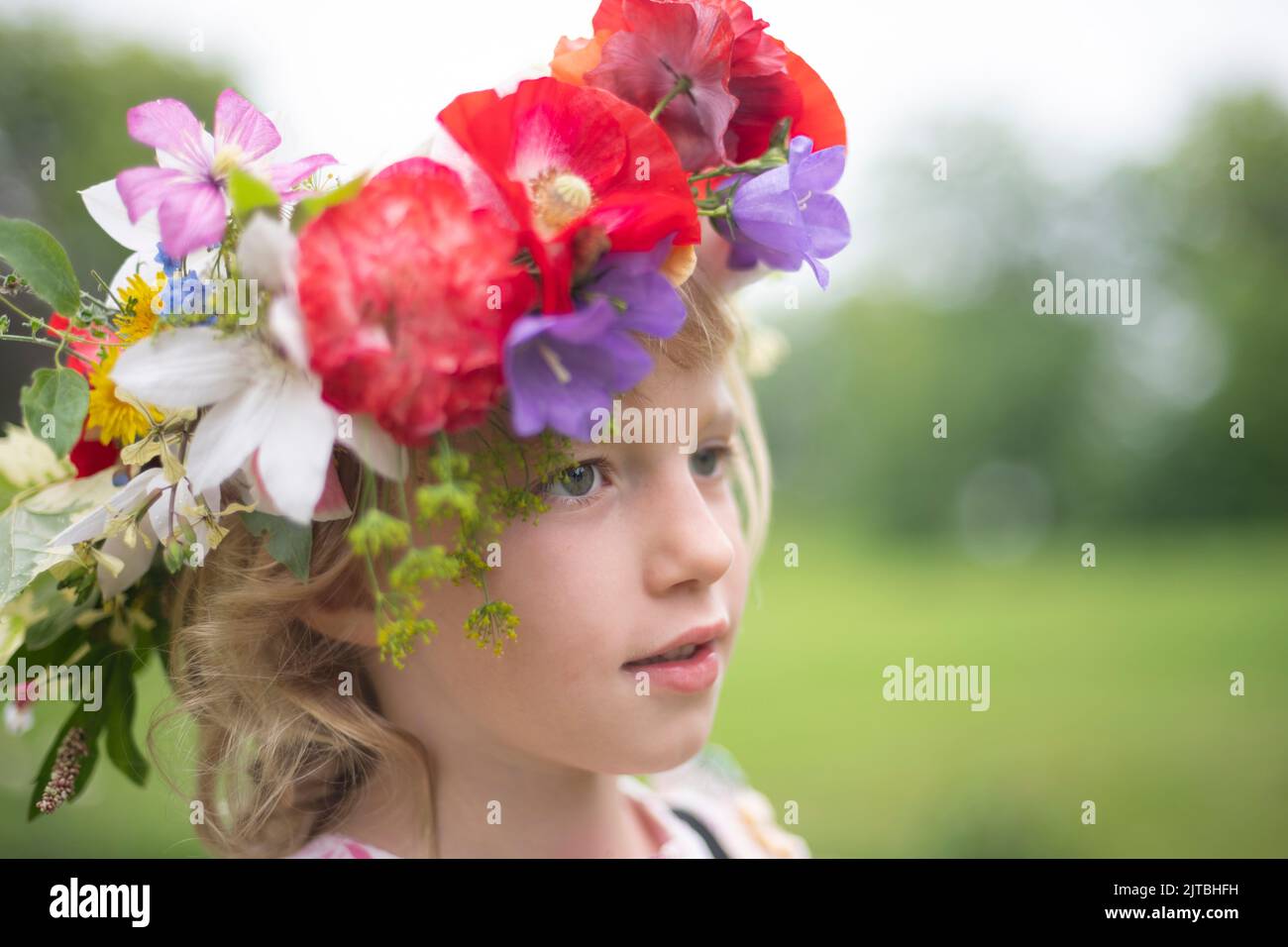 A young girl with blonde hair wears a flower crown with red poppies, blue campanula, and leaves against the green backdrop of a summer day. Stock Photo