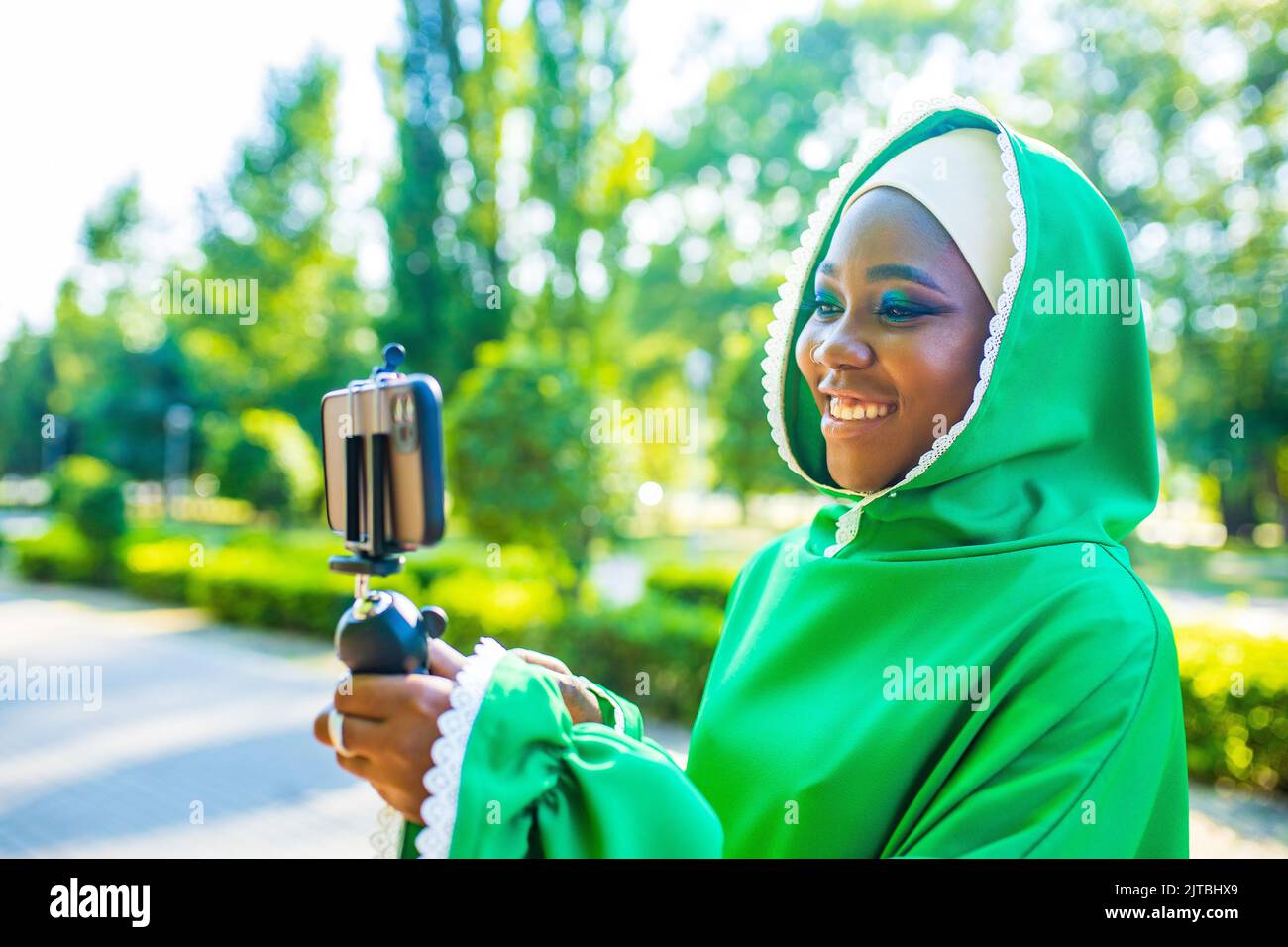 latin hispanic wonan in green muslim hijab with bright make up and nose piercing taking video stream outdoors summer park Stock Photo
