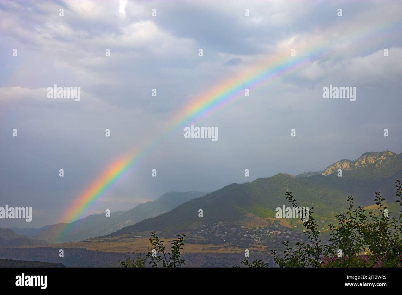 Rainbow and sun rays on a cloudy and rainy day, with a green forest, mountains and a village in the background, in summer Stock Photo