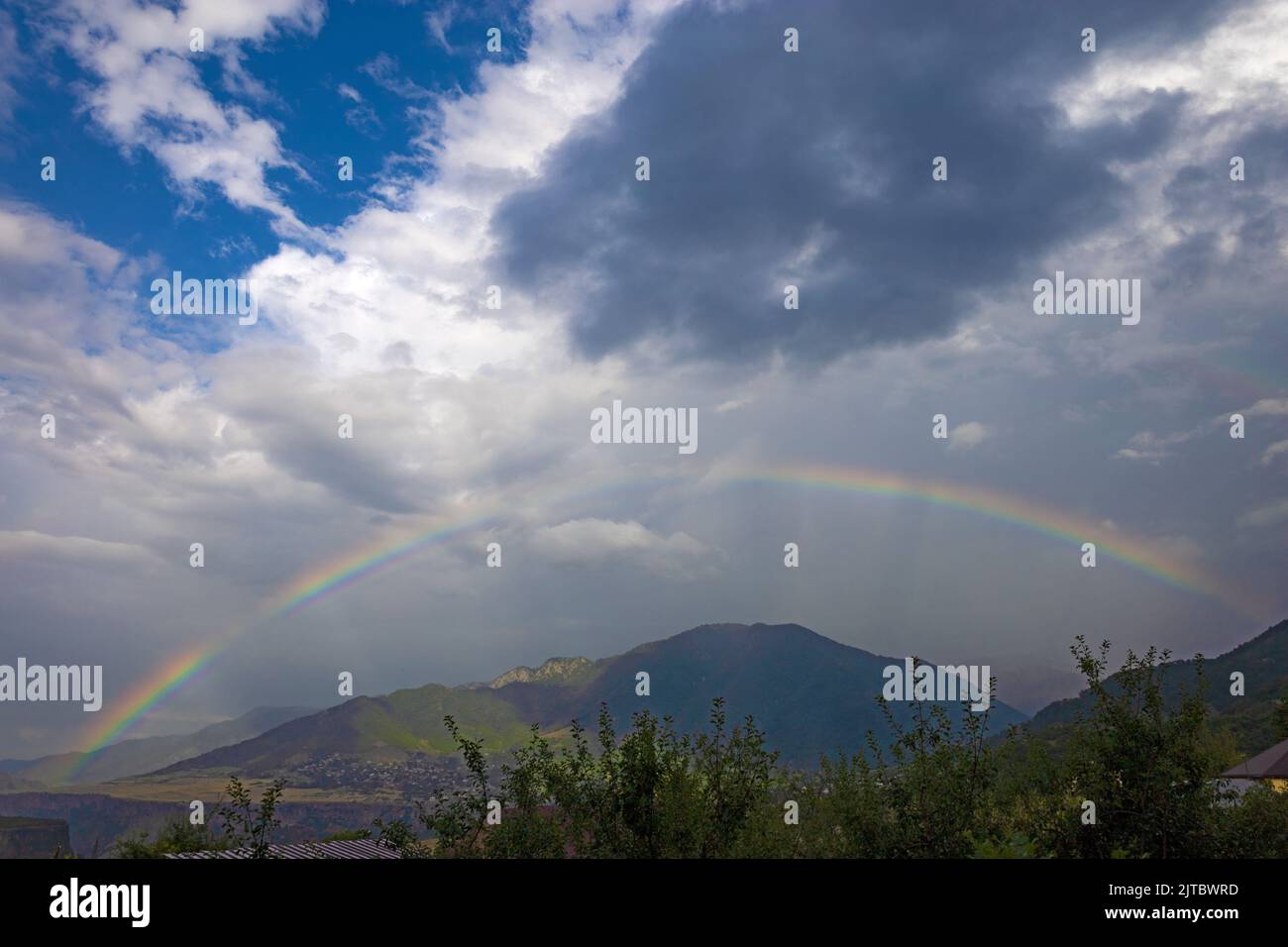 Full complete rainbow and sun rays on a cloudy and rainy day, with a green forest, mountains and a village in the background, in summer Stock Photo