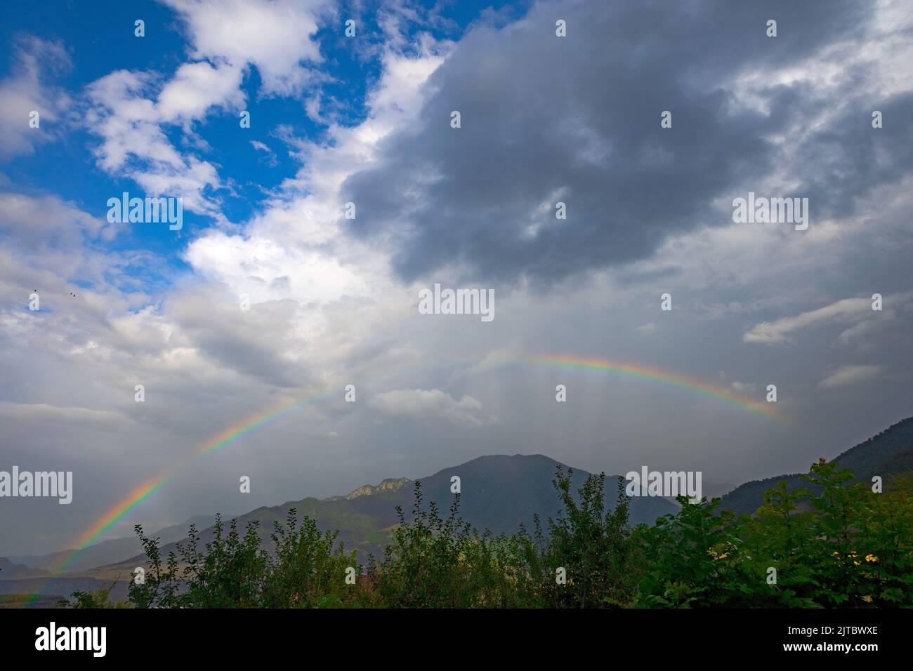 Full complete rainbow and sun rays on a cloudy and rainy day, with a green forest, mountains and a village in the background, in summer Stock Photo