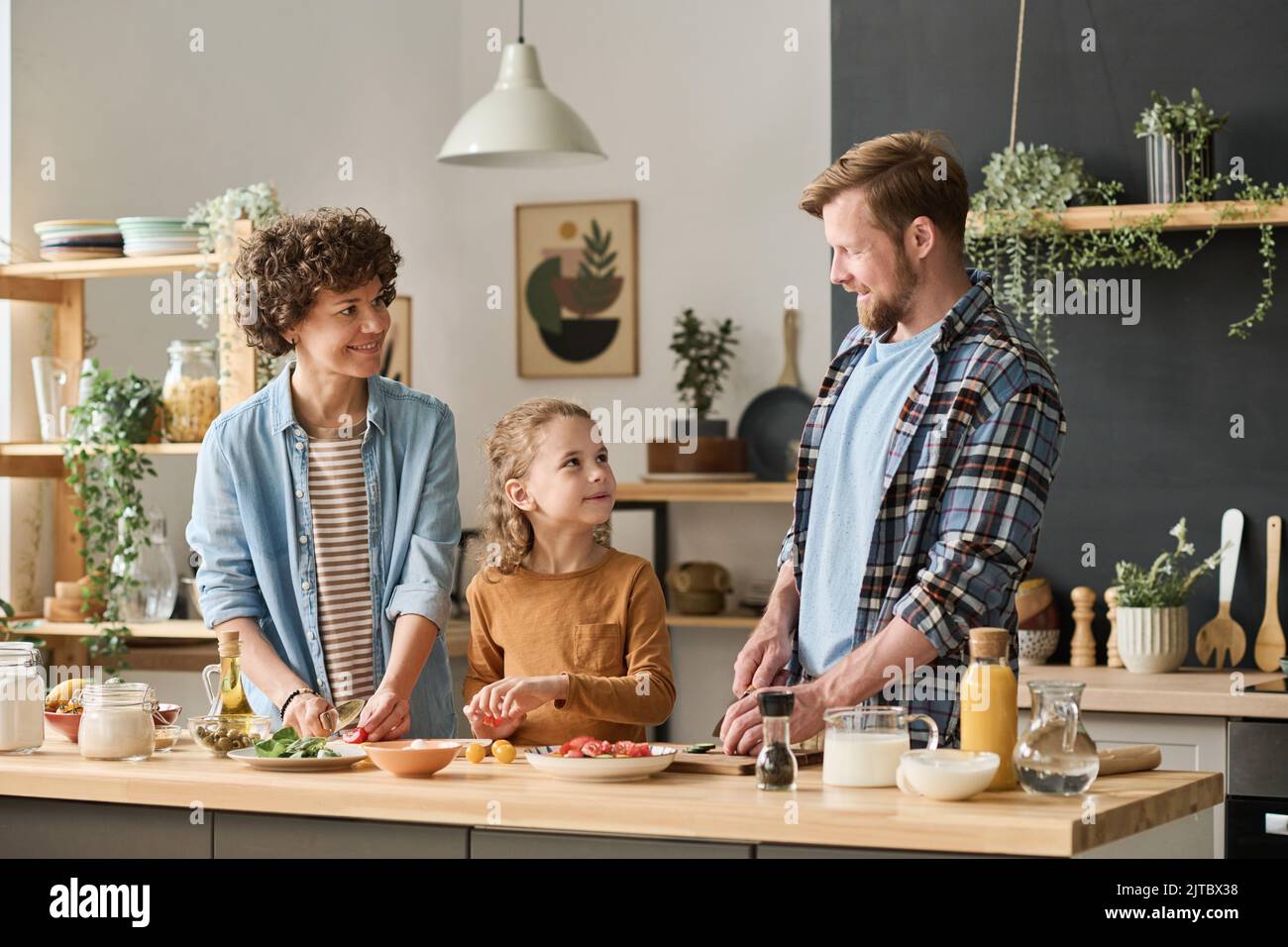 Family of three cutting vegetables for salad at table, they preparing dinner together at home with their little son Stock Photo
