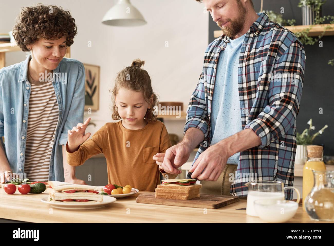 Little boy helping his parents to cook sandwiches with vegetables at table in domestic kitchen Stock Photo