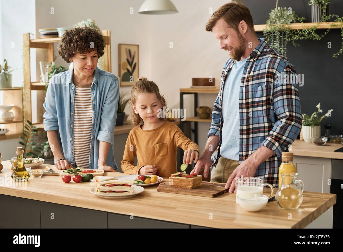 Little boy putting fresh vegetables on bread to cook sandwiches together with his parents in kitchen Stock Photo